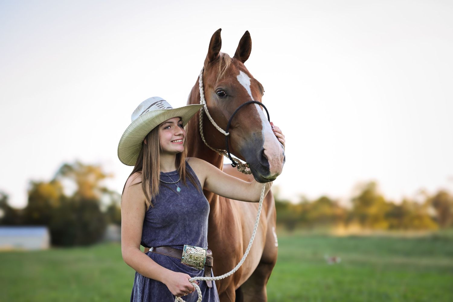 A chestnut horse stands with its handler in a blue dress and cowboy hat in a sunlit pasture during golden hour.