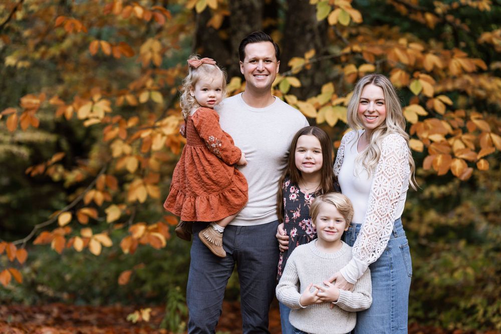 A family of five poses together outdoors against a backdrop of autumn foliage with orange and yellow leaves.