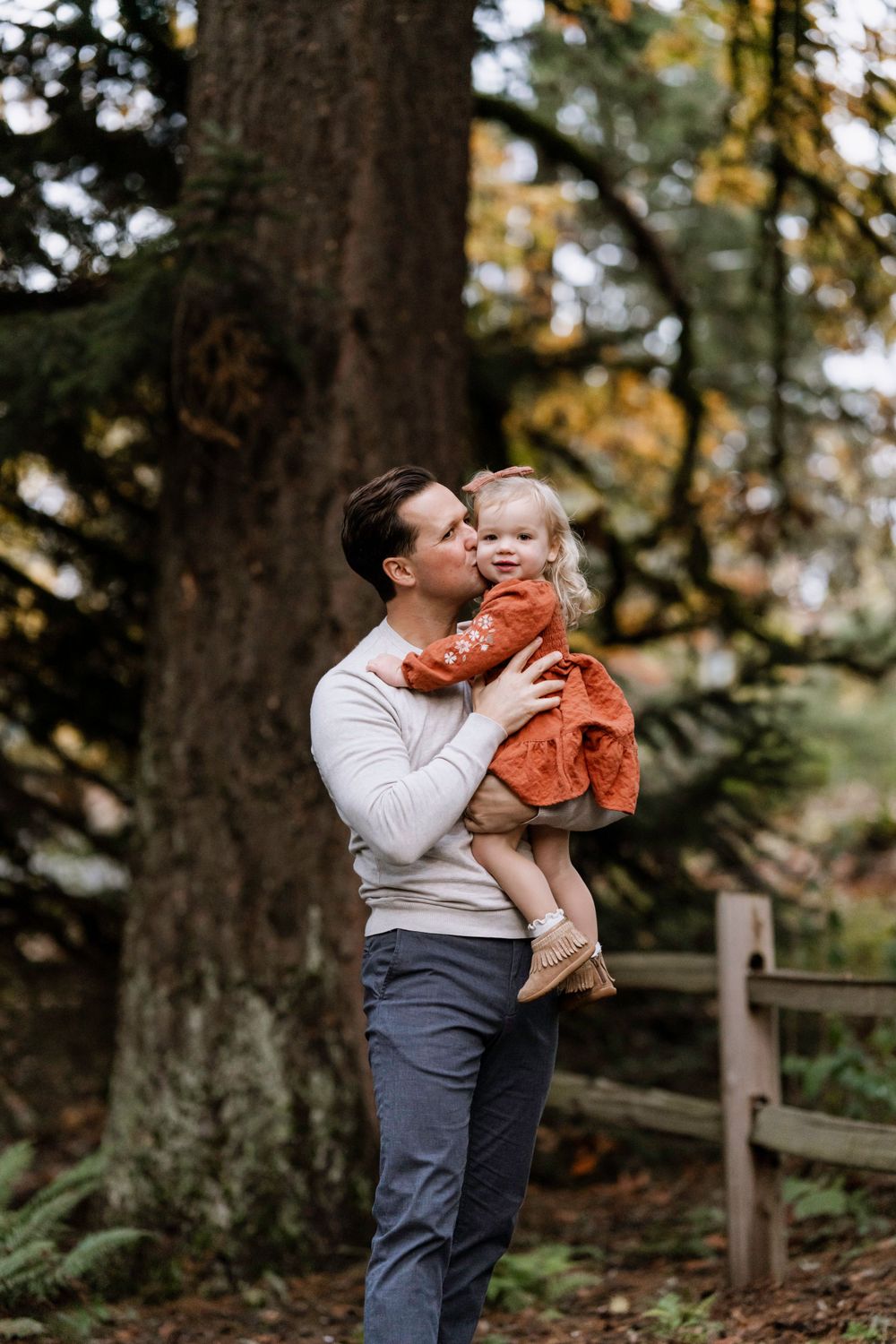 An adult holds a small child wearing a bright orange dress in a wooded outdoor setting during autumn.