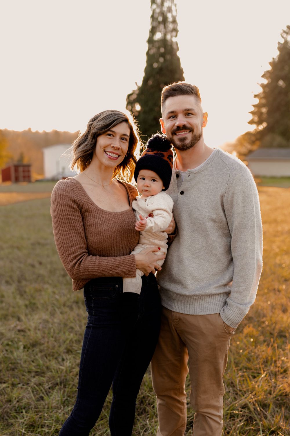 Family of three poses together outdoors at sunset in a grassy field during golden hour.