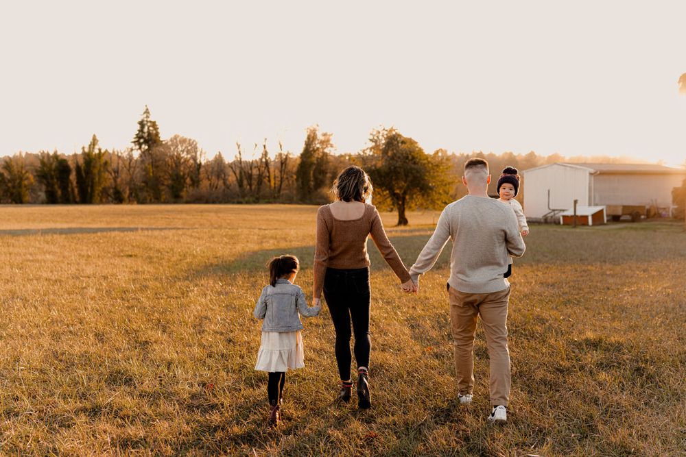 A family walks hand in hand across a golden field at sunset with tall trees in the background. Family photography Fleck of Light.