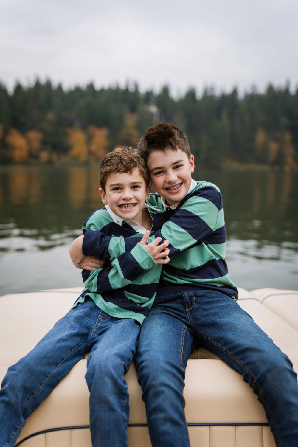 Two children in matching striped shirts sitting together on a boat with an autumn lake and forest backdrop.