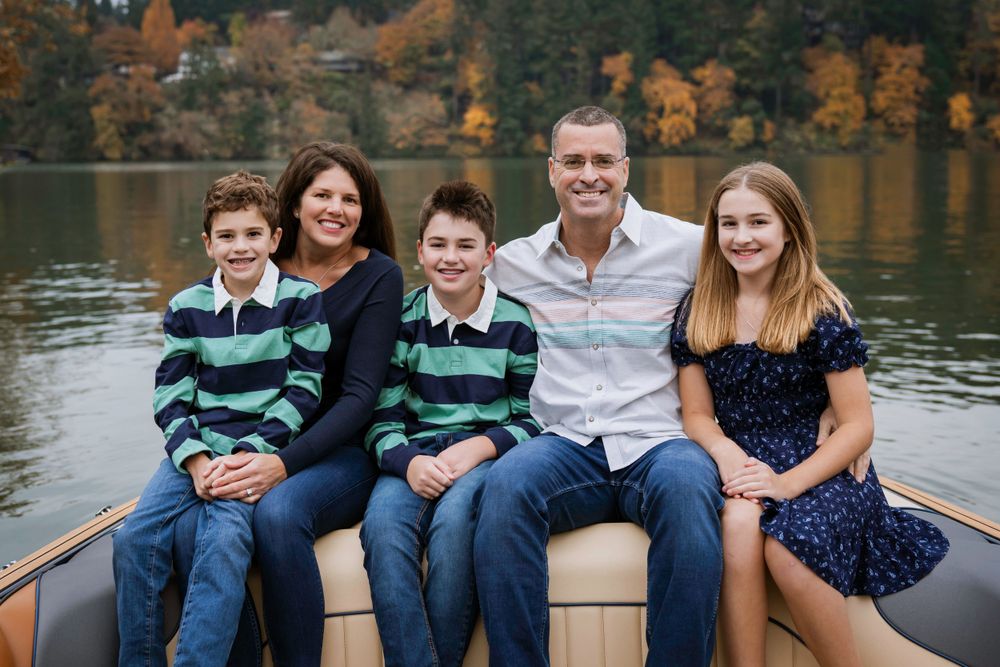 A group sits together on a boat bench with a scenic autumn lake and colorful foliage in the background. Fleck of Light Photography.