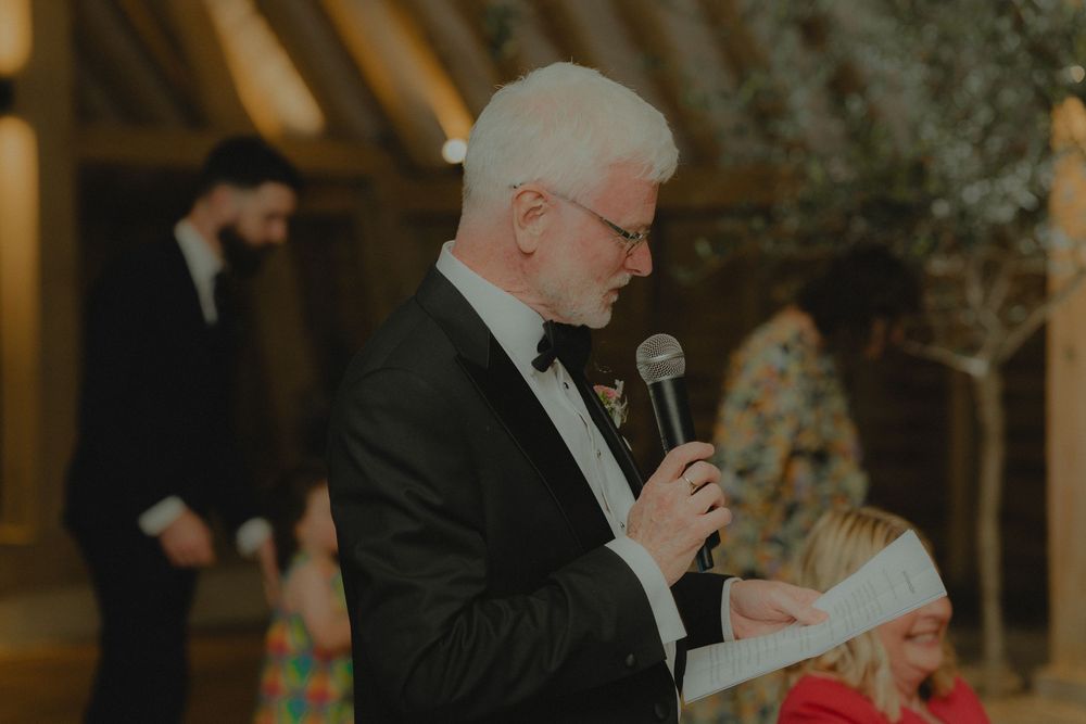 Elderly man in tuxedo delivering wedding speech into microphone at elegant barn venue.