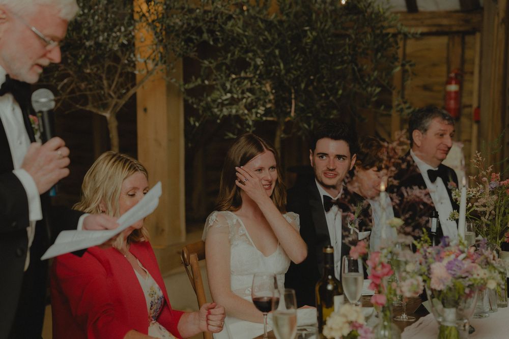 Bride wipes tears during emotional wedding speech at rustic barn reception with floral centerpieces.