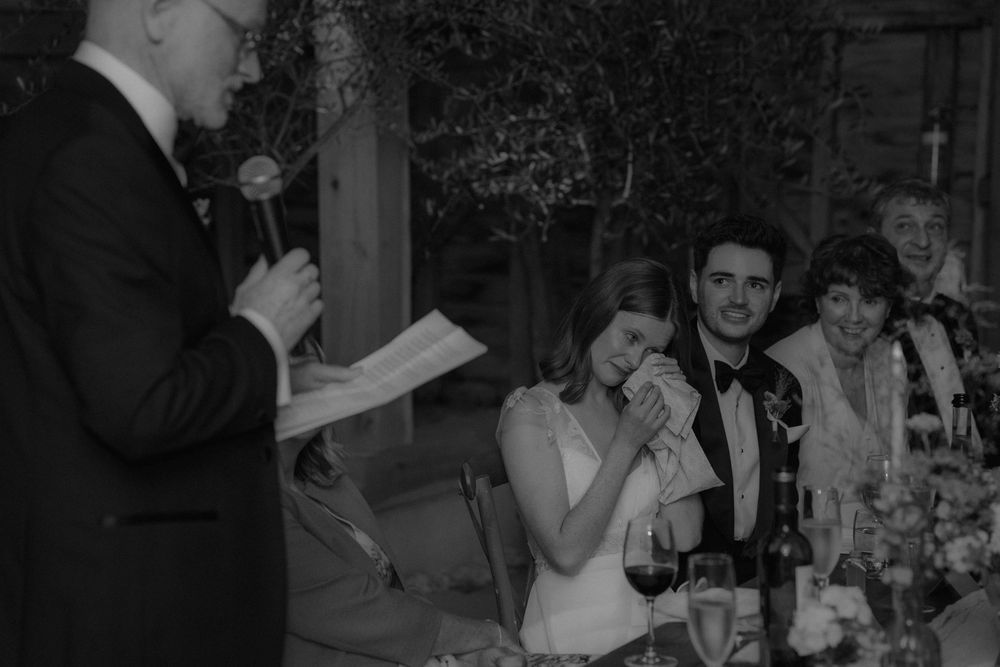Bride wipes tears during emotional wedding speech while groom smiles beside her at reception dinner.