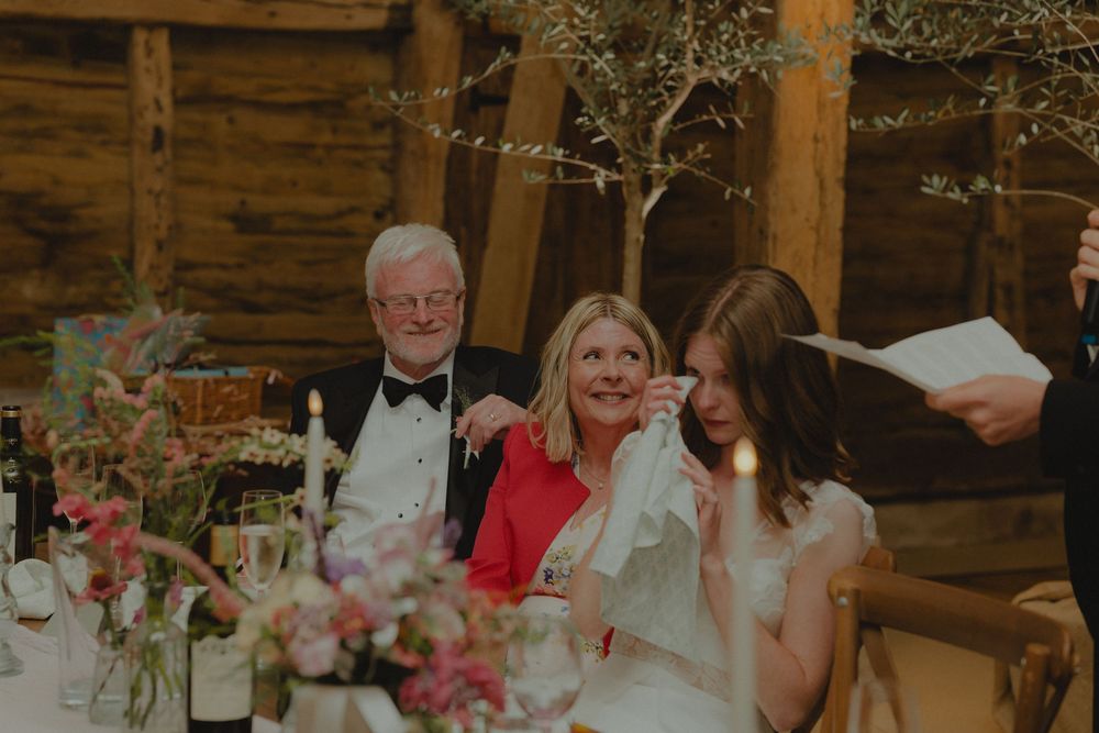 Father of bride poses with emotional bride and wedding guest at barn reception table.