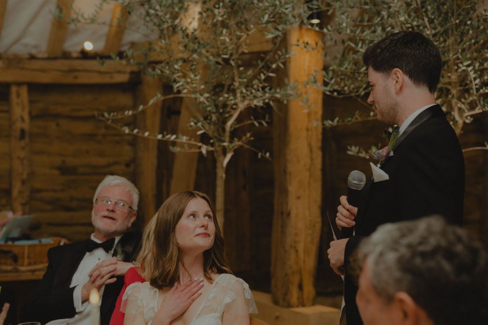 Groom delivers wedding speech at rustic barn reception while bride and elderly guest listen attentively.
