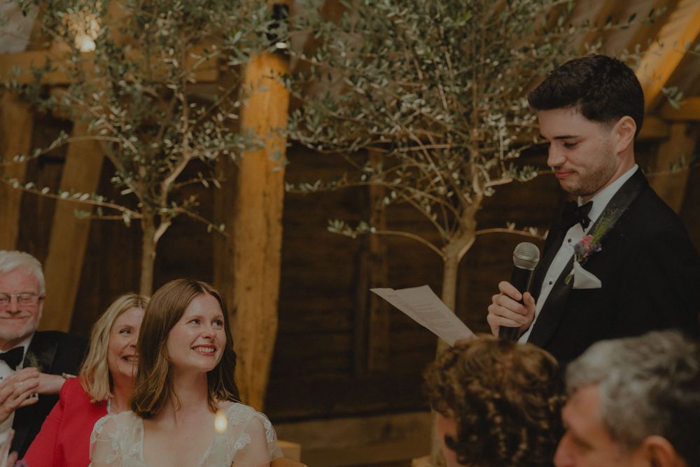 Groom delivers heartfelt wedding speech as bride smiles at rustic barn venue with fairy lights.