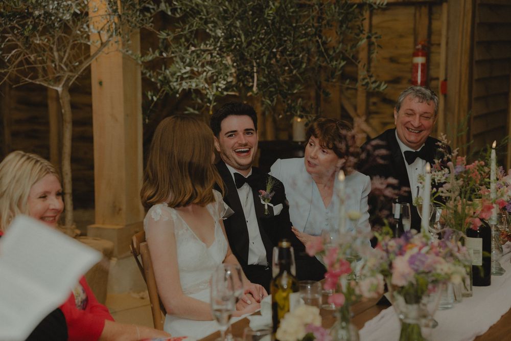 Bride and groom laughing joyfully at wedding reception dinner table with guests and floral centerpieces.