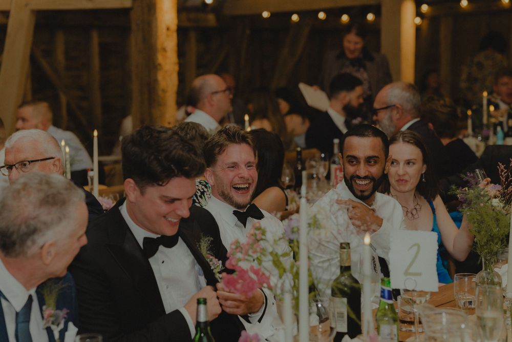 Guests laughing and celebrating at a wedding reception dinner inside a rustic barn venue with string lights.