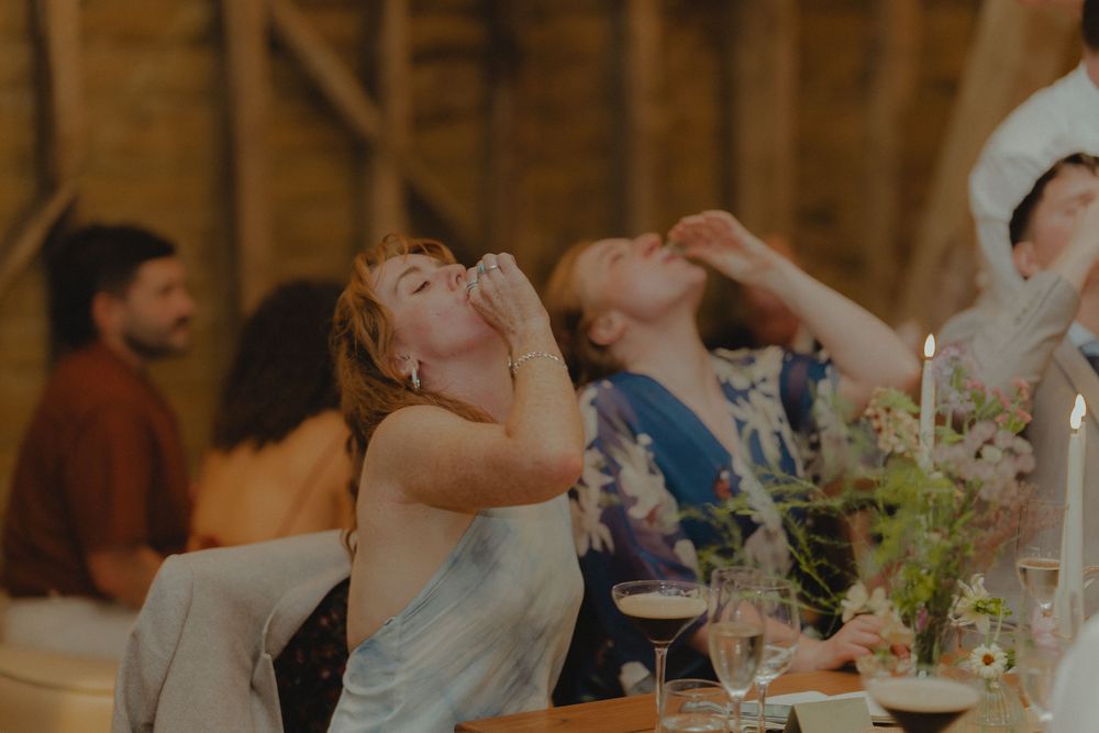 Wedding guests laughing and drinking at rustic barn reception dinner table with candles and flowers.
