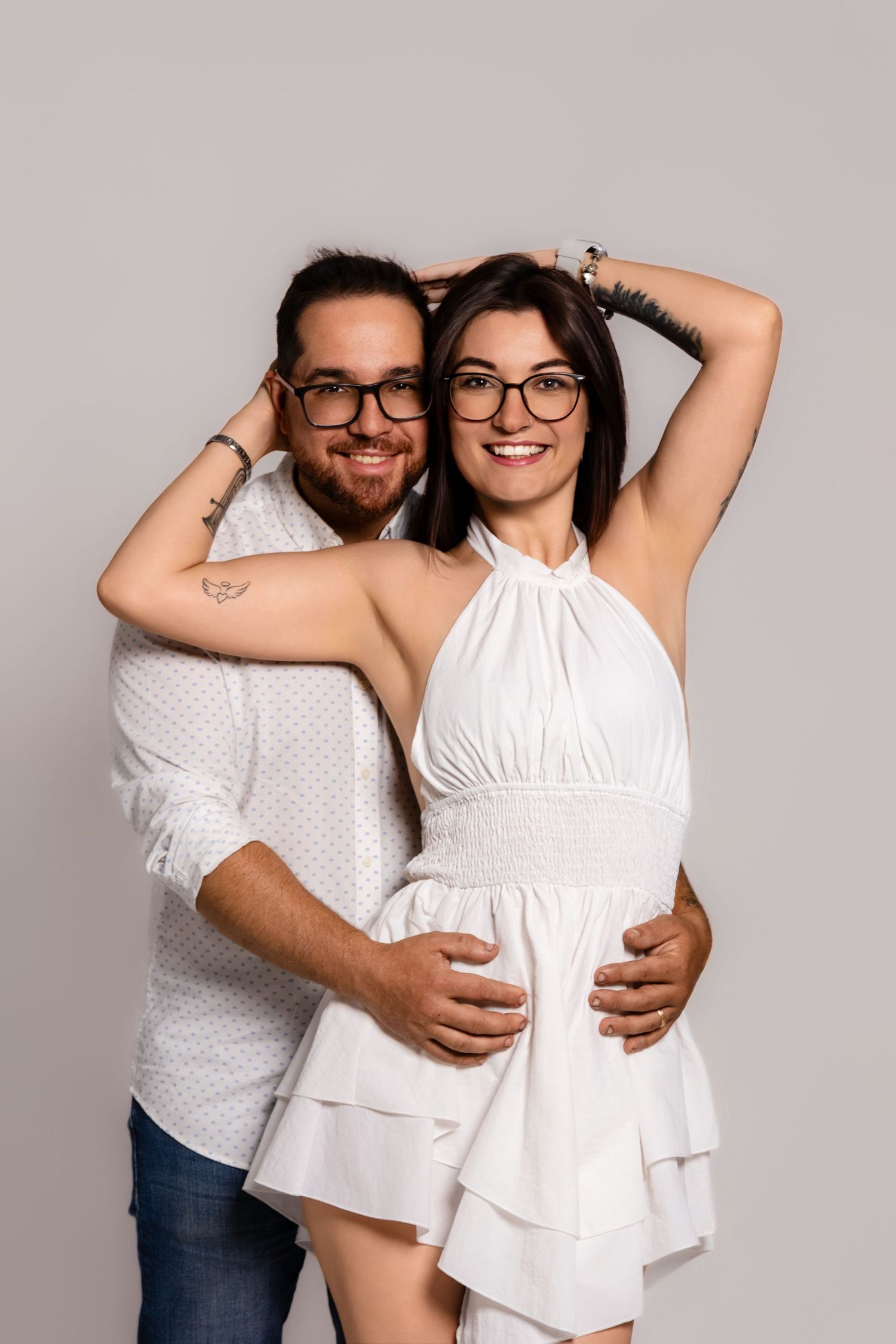 Playful couple in white outfits poses together against light grey backdrop.