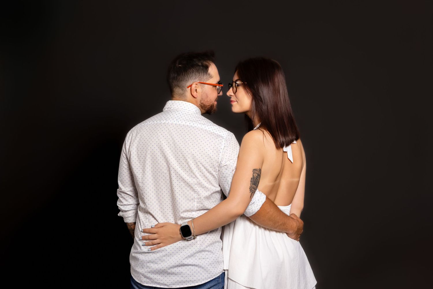 Romantic couple in white clothing embraces against dark studio backdrop.