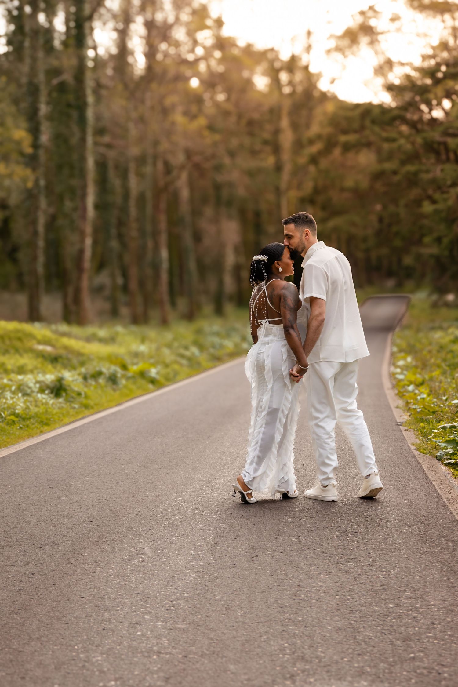 A couple dressed in white walks together on a tree-lined path during golden hour.