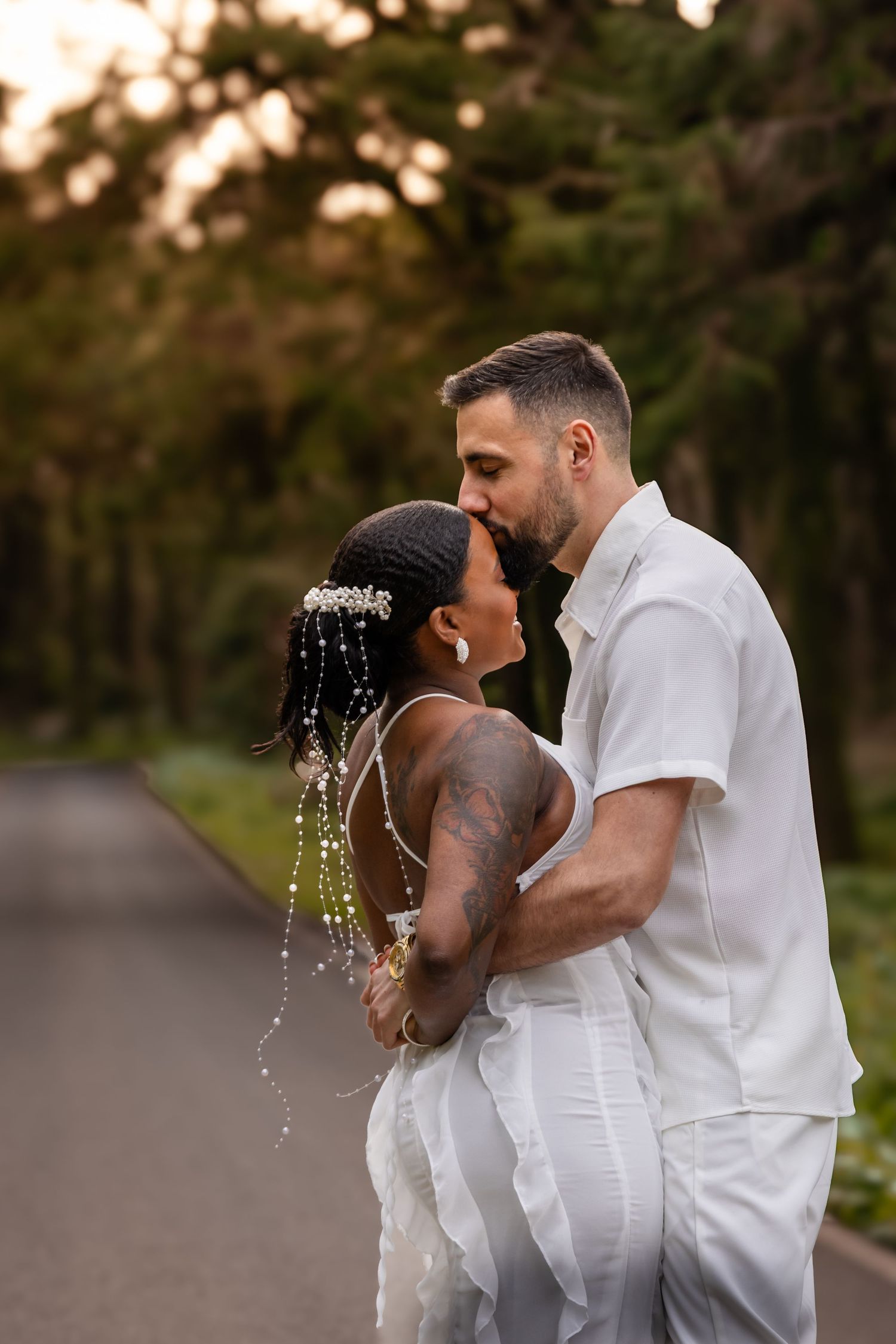 A couple in white outfits shares a playful moment with water splashing on a wooded path at sunset.