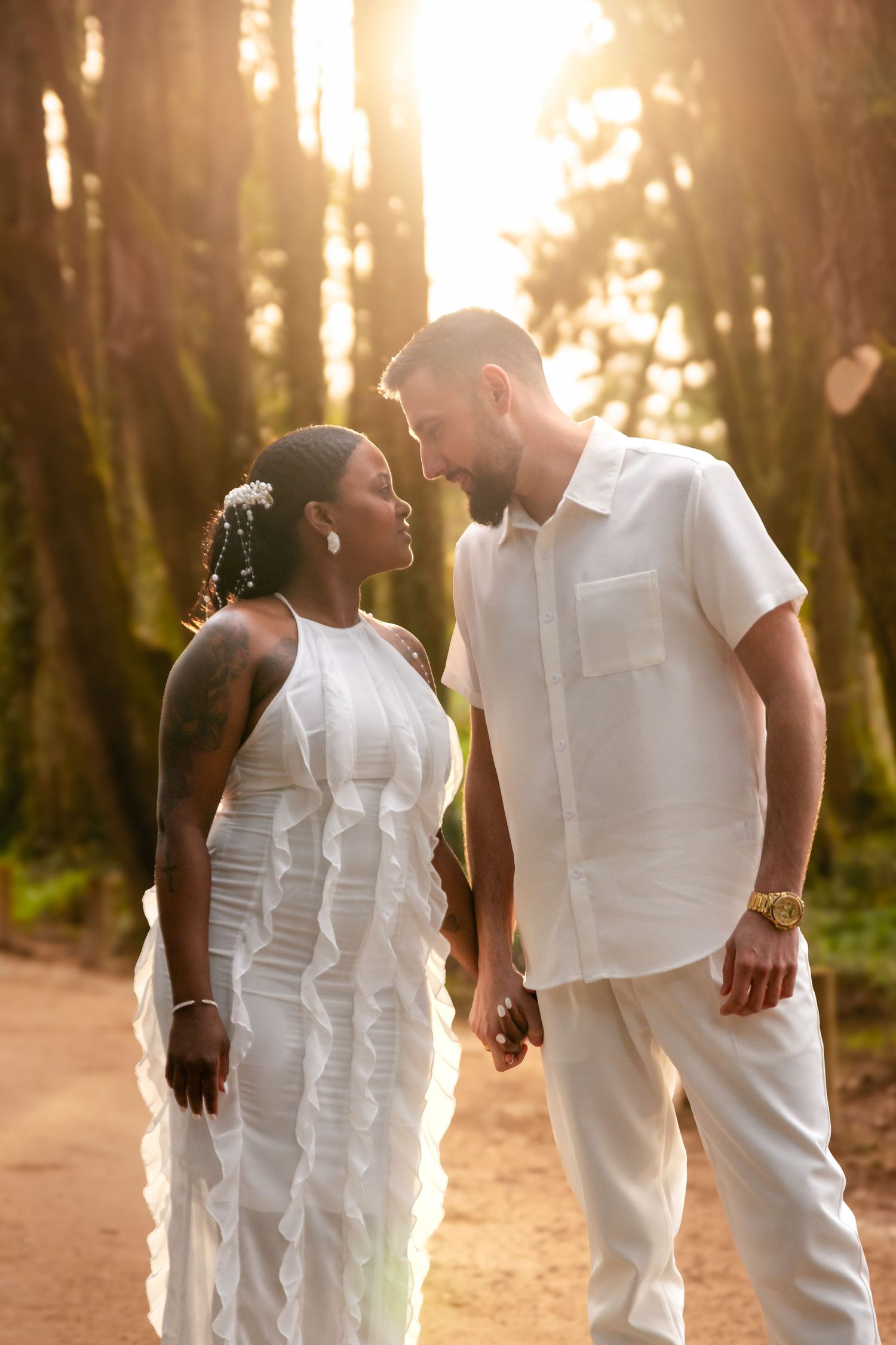 A romantic couple in matching white outfits walks hand in hand on a sunlit forest trail.