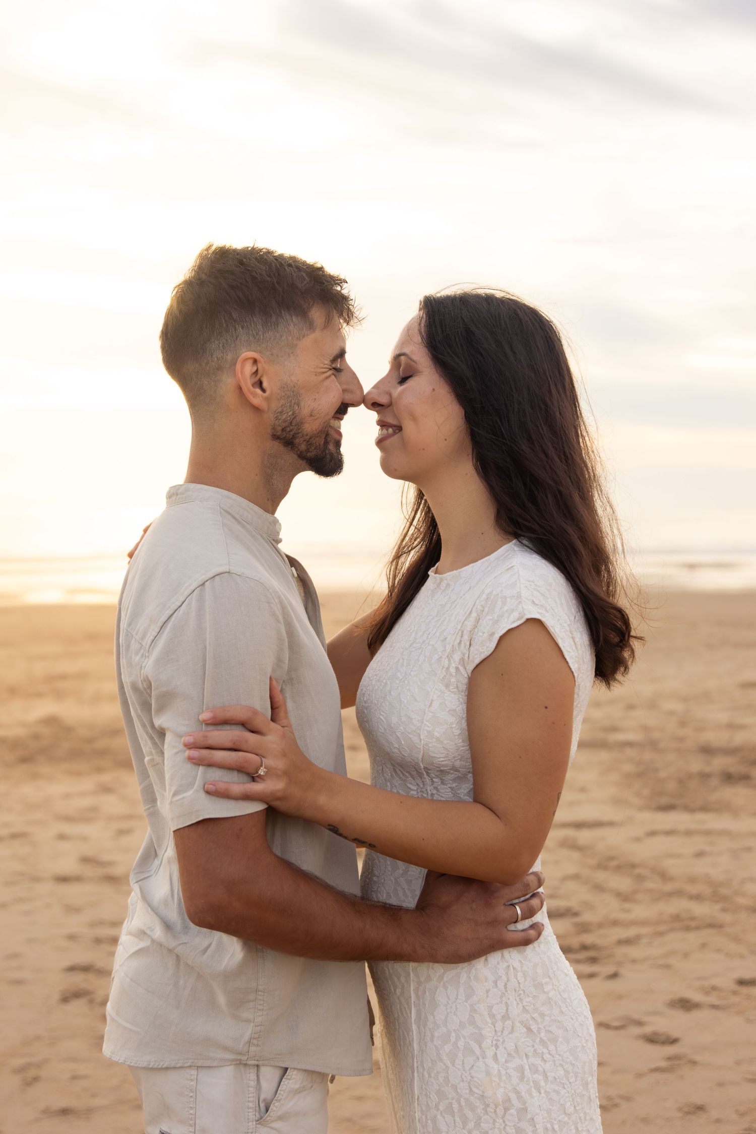 Intimate moment between couple in white outfits embracing on sandy beach at sunset.