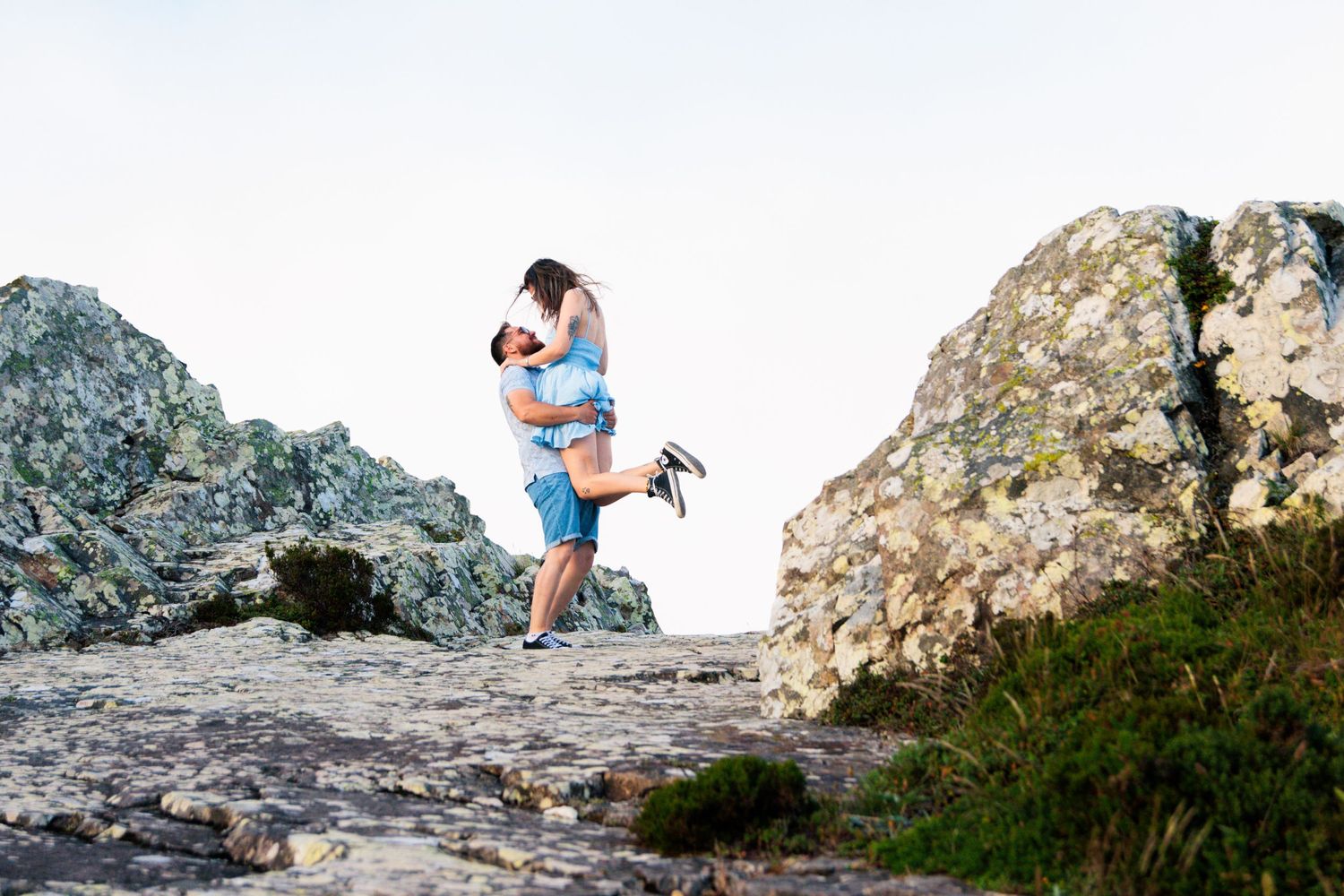 A couple embraces on rocky cliffs overlooking a scenic landscape.