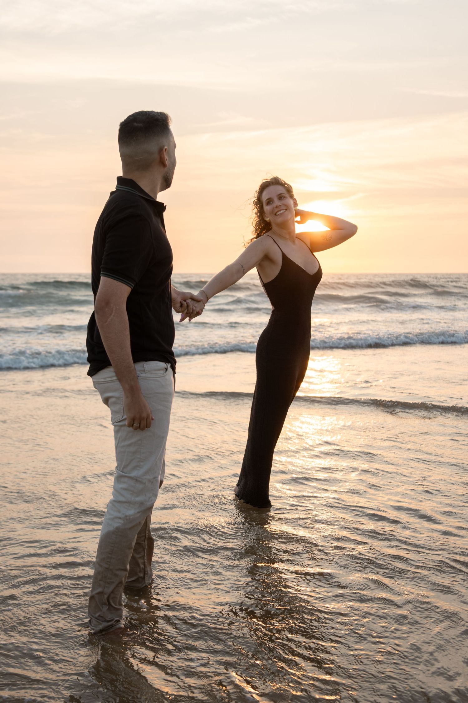 Couple standing in ocean waves during golden sunset at beach creating romantic silhouettes.