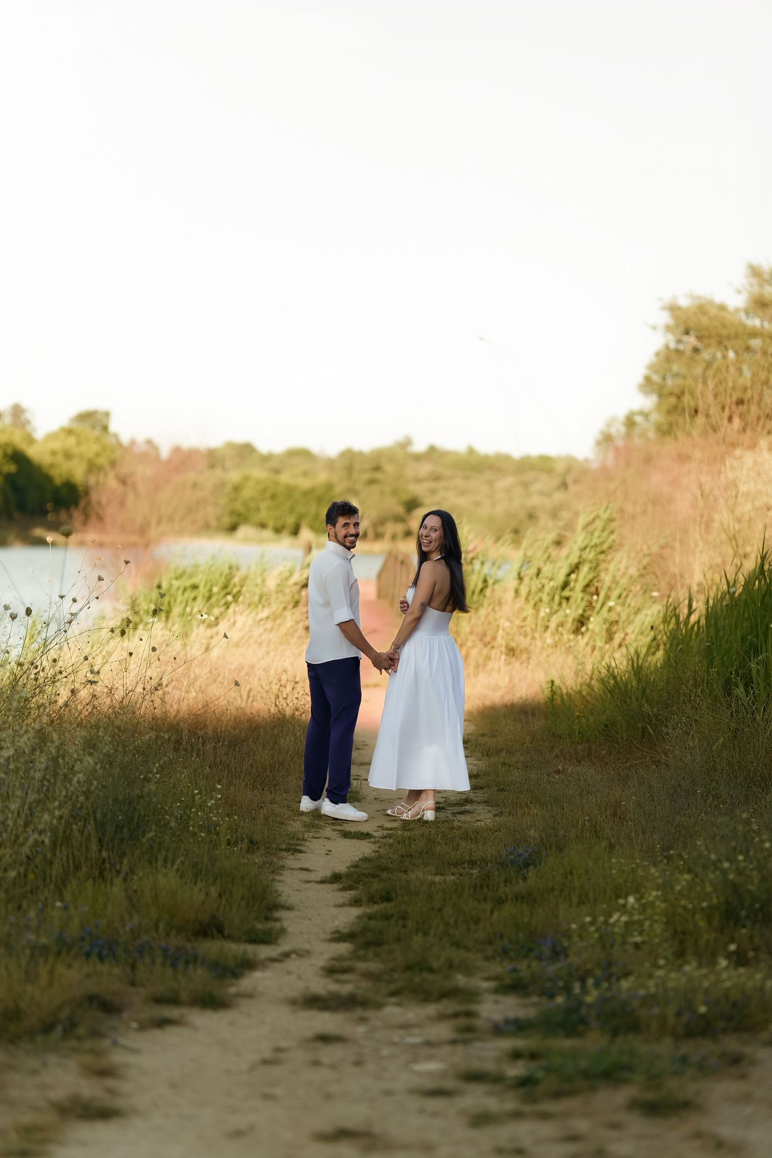 Couple holding hands on a scenic nature path at golden hour, woman in white dress, man in casual attire.