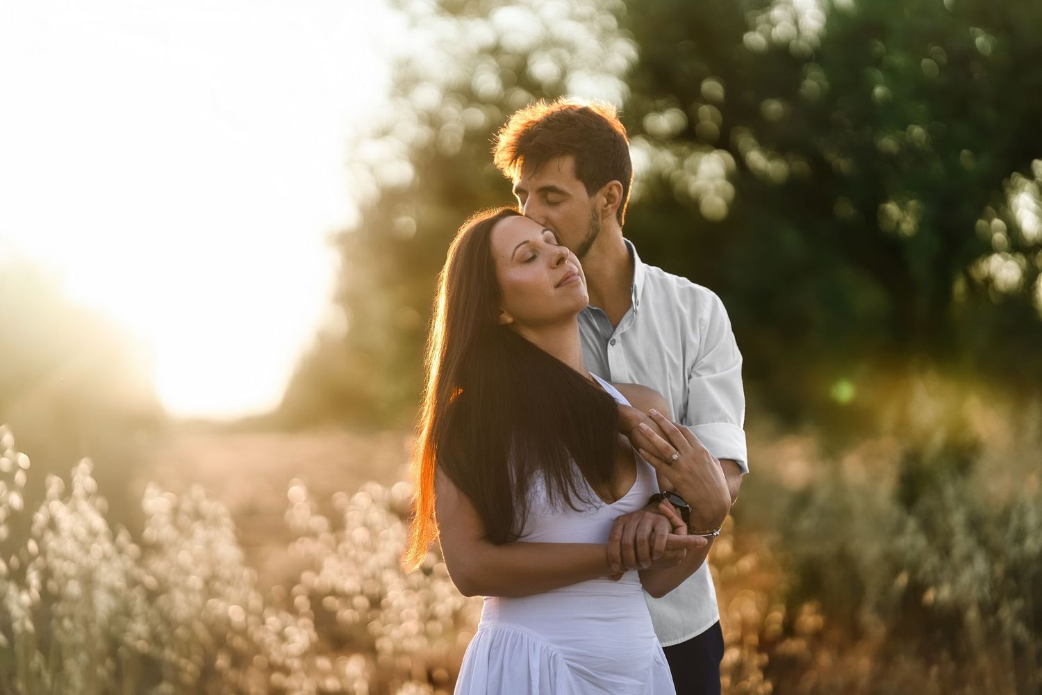 Romantic couple embracing in a sunlit field at golden hour, woman in white dress leaning into man's kiss.