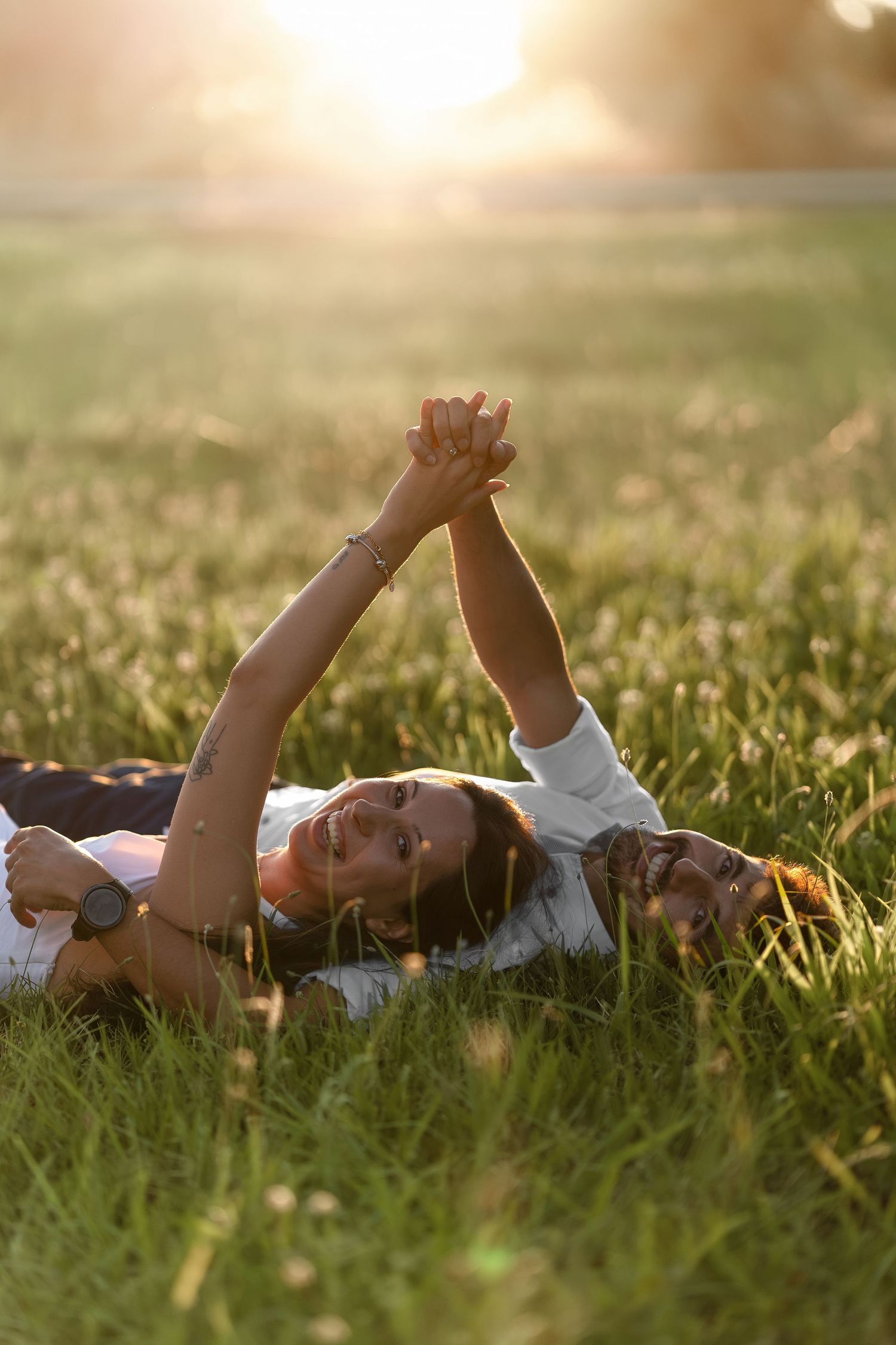Two people lying on green grass holding hands raised up, bathed in warm golden sunset light in a field.