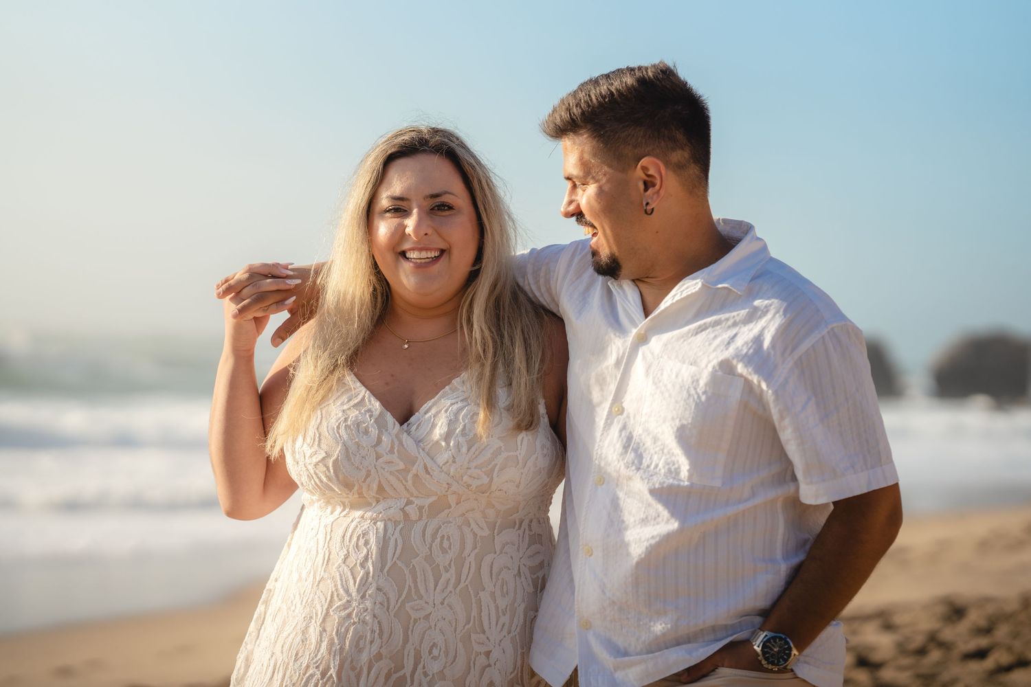 A smiling couple poses together on a sunny beach, woman in white lace dress, man in white shirt, ocean waves behind them.