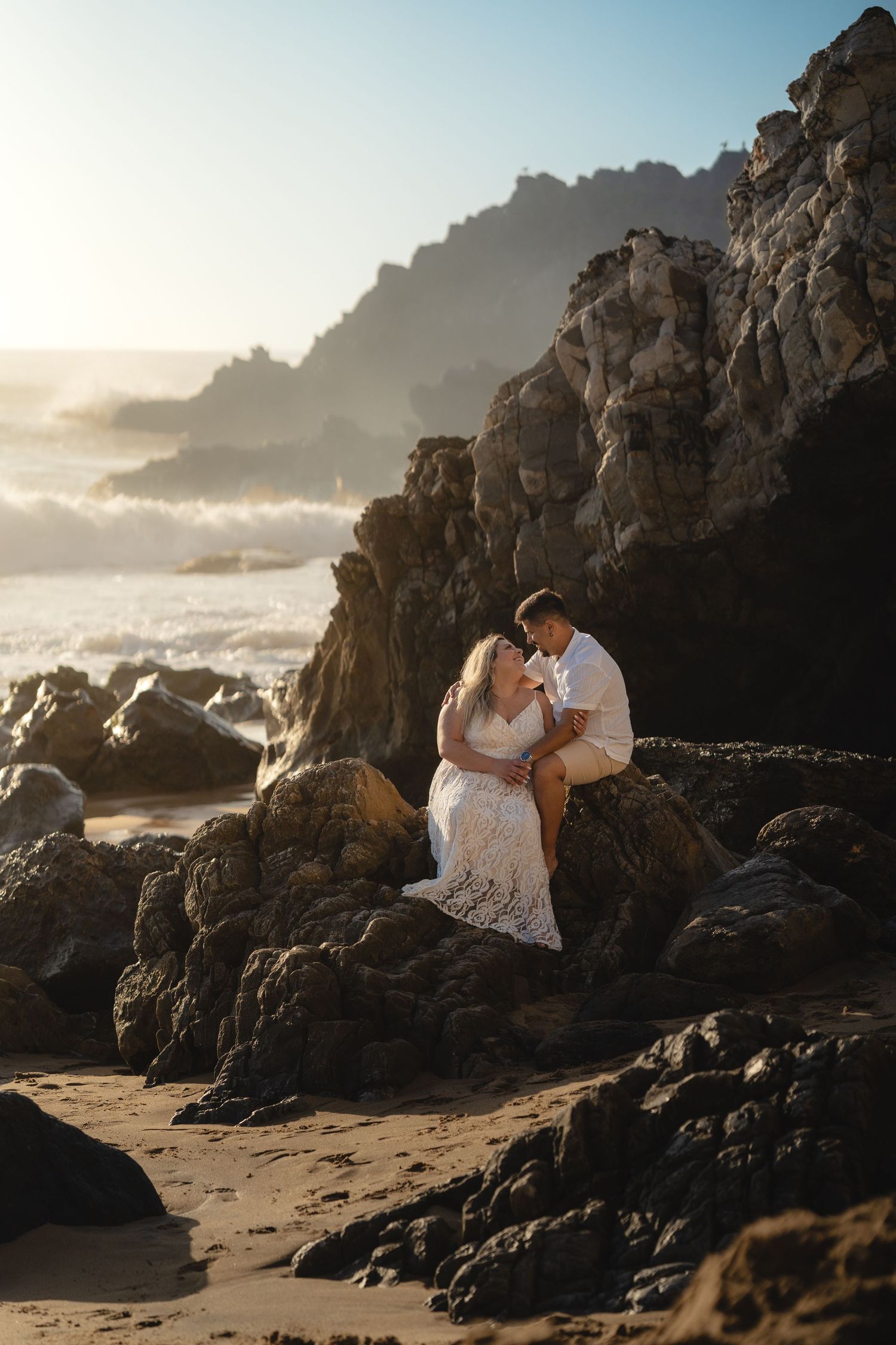 Couple sharing a romantic moment on coastal rocks at sunset, waves crashing behind them on a scenic beach.