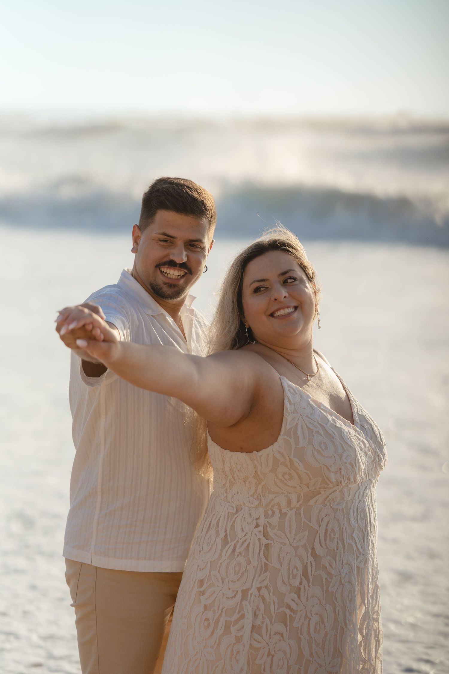 A smiling couple dances together on a sunny beach, dressed in white and beige outfits with ocean waves behind them.