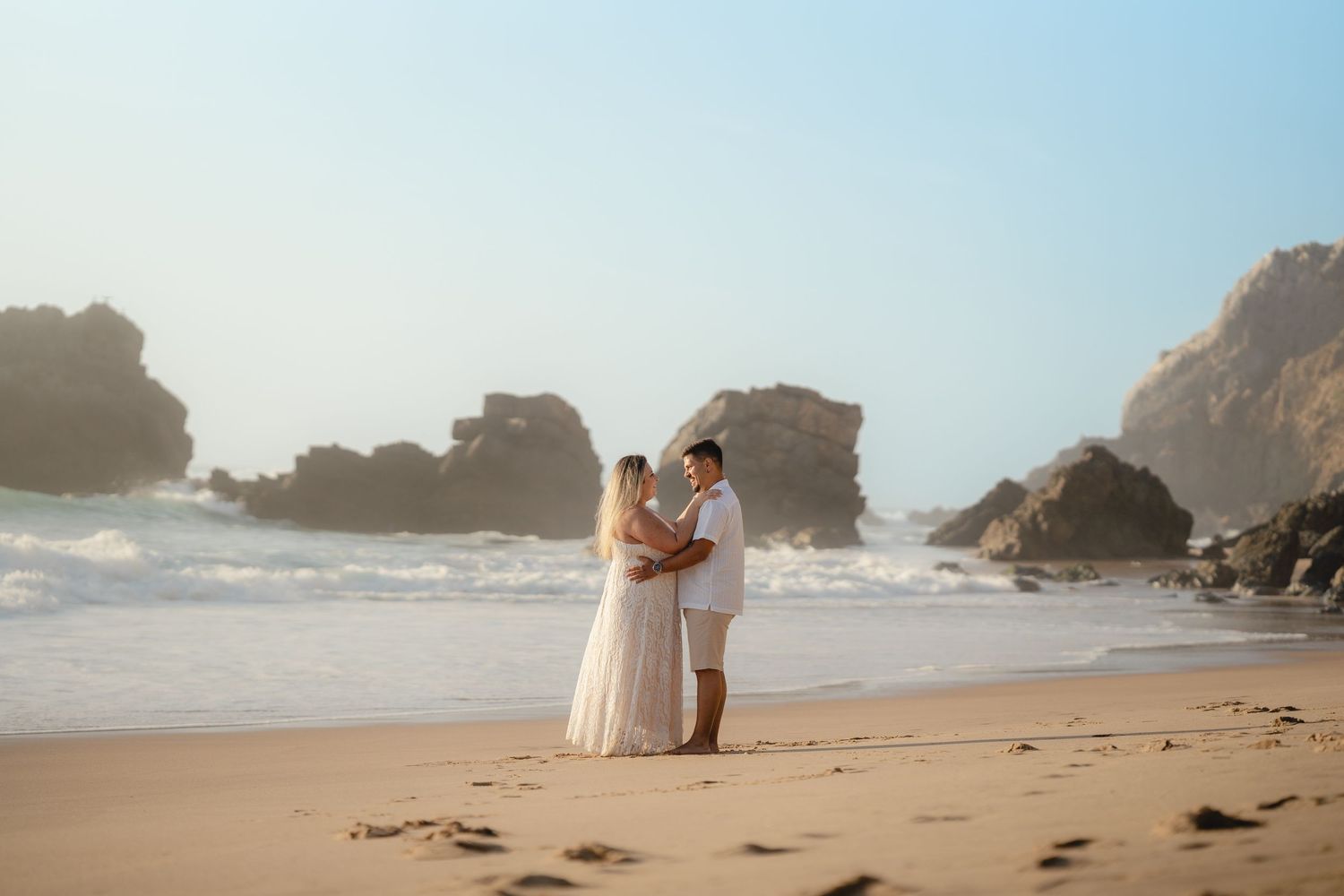 A couple in white attire holds hands on a sandy beach with rocky cliffs and ocean waves behind them.