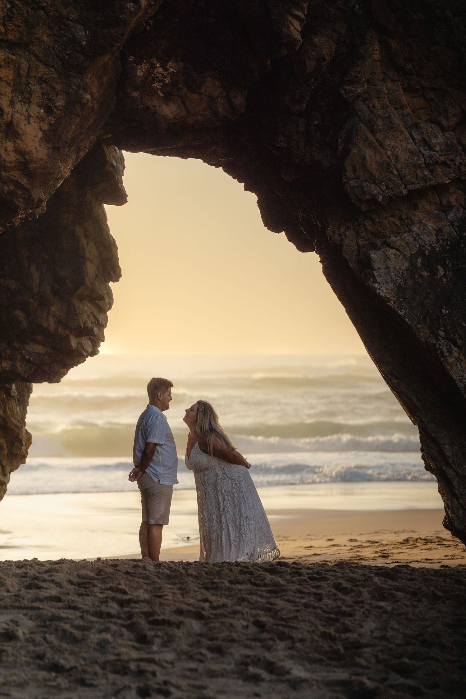 Romantic couple standing face to face under a natural rock arch on a sandy beach at golden sunset with ocean waves.