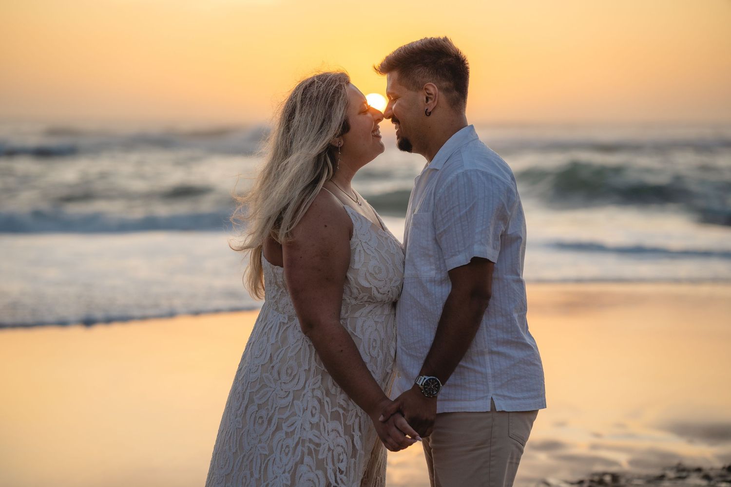 Couple sharing a romantic kiss at sunset on the beach, holding hands in wedding attire by ocean waves.