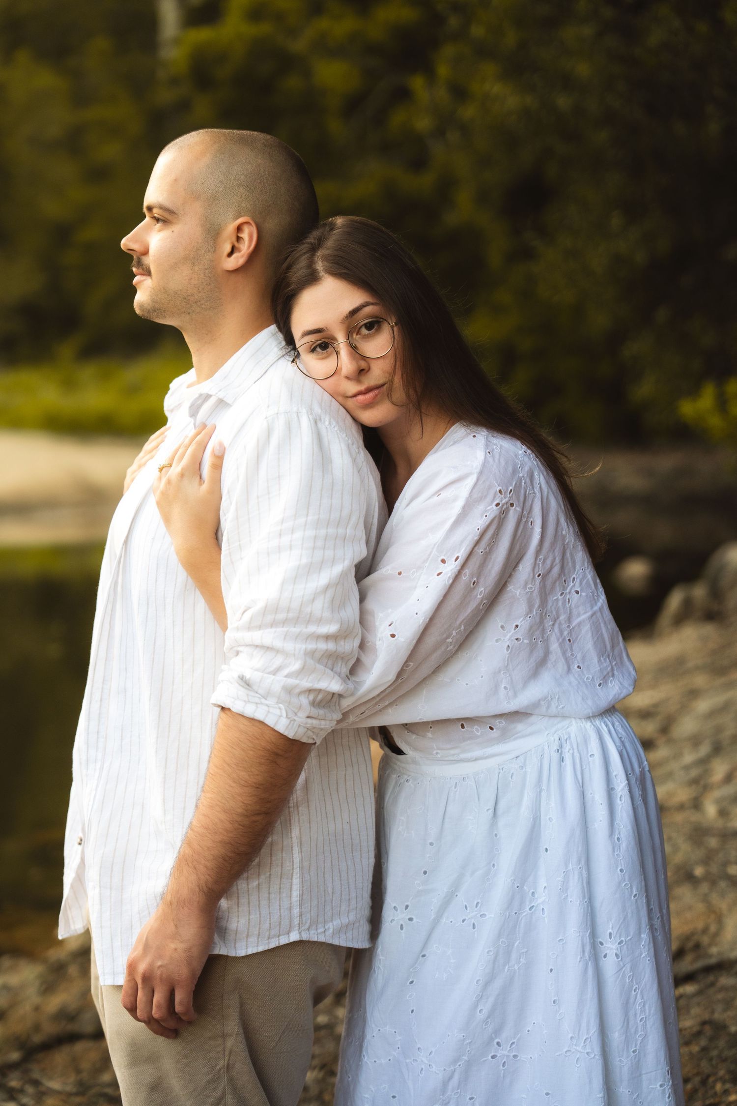 Romantic couple in white outfits embrace by a scenic lakeside during a photo session.