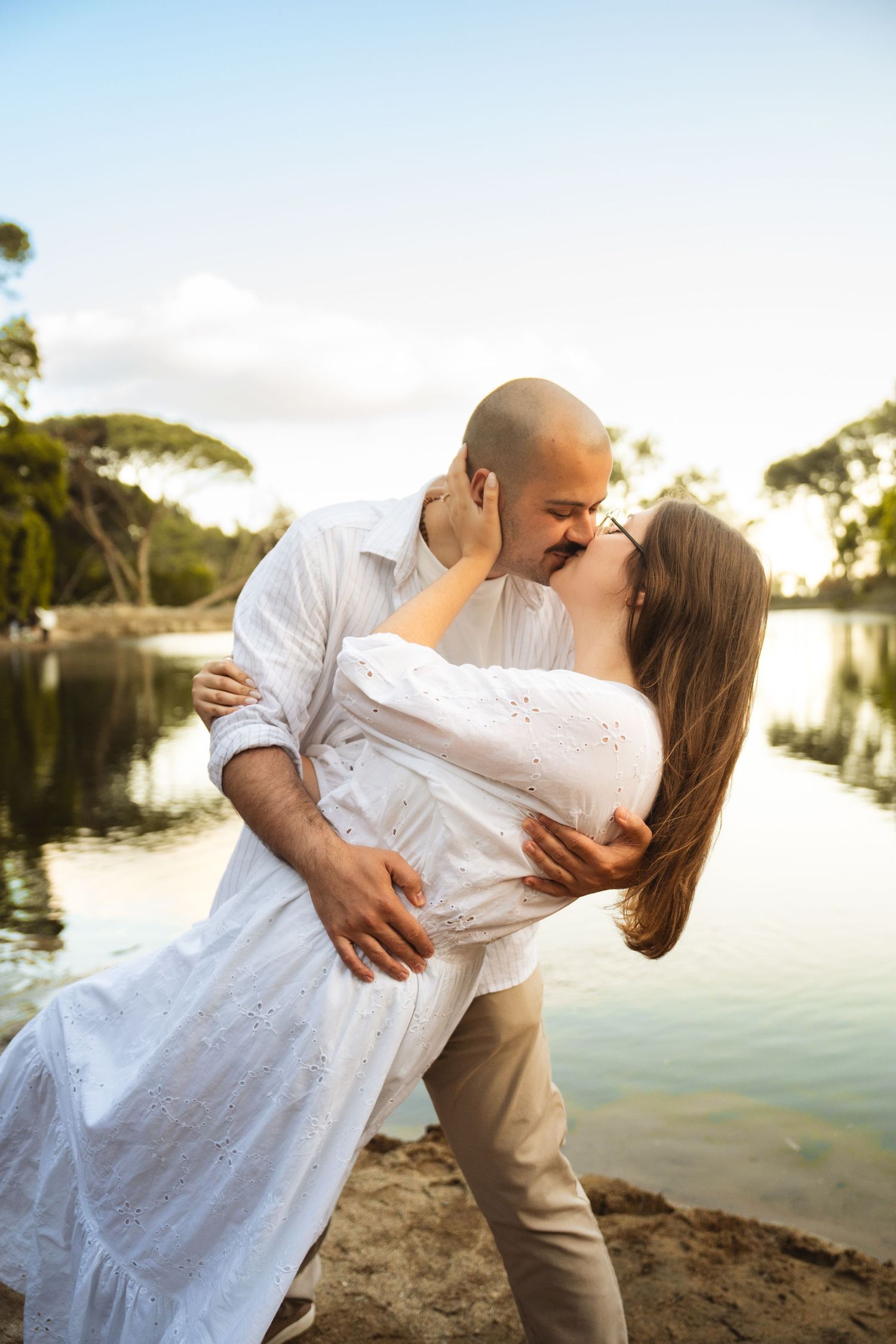Couple in white clothing share a romantic kiss by a serene lake at sunset.
