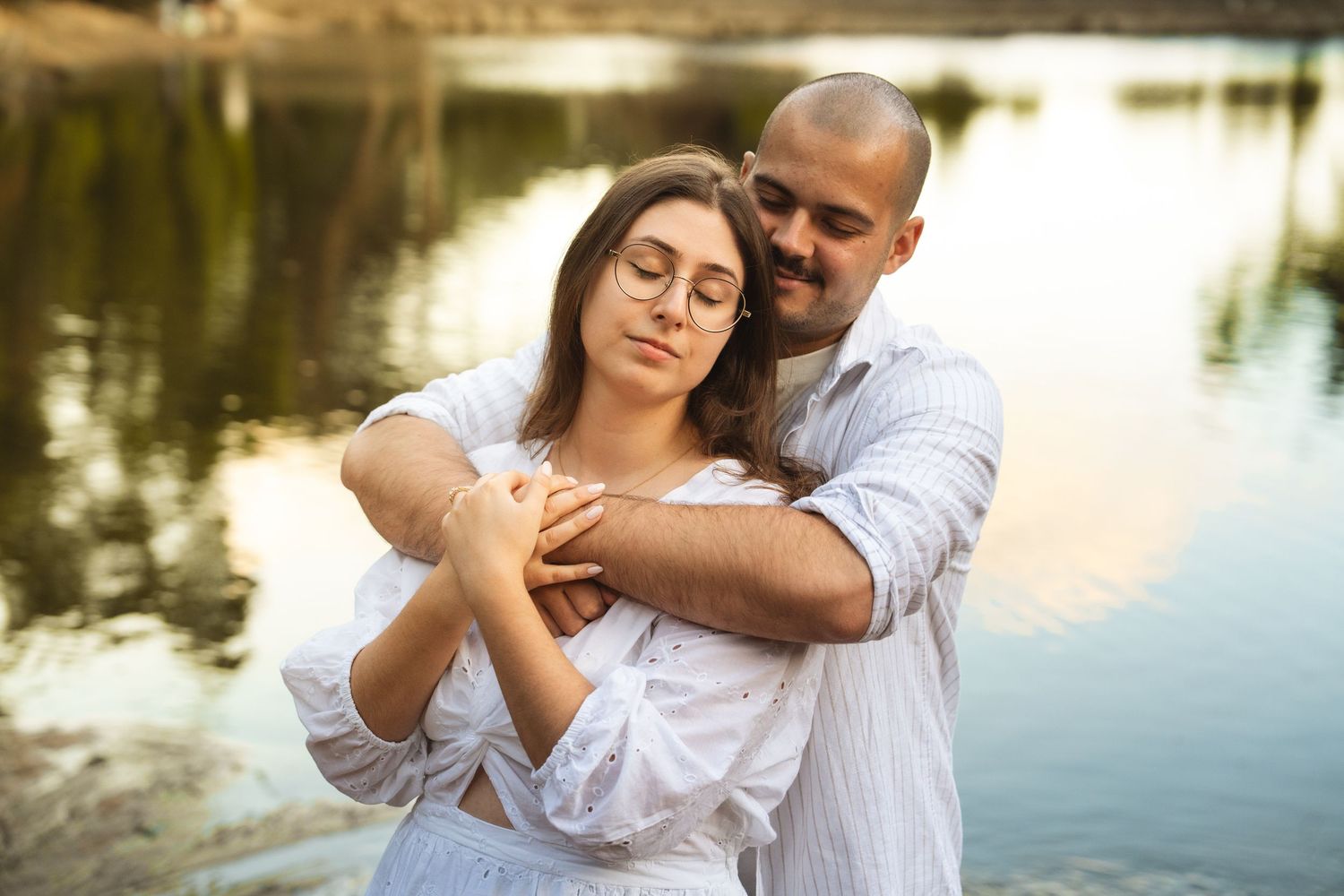 Couple in white clothing embrace tenderly by a tranquil lake.