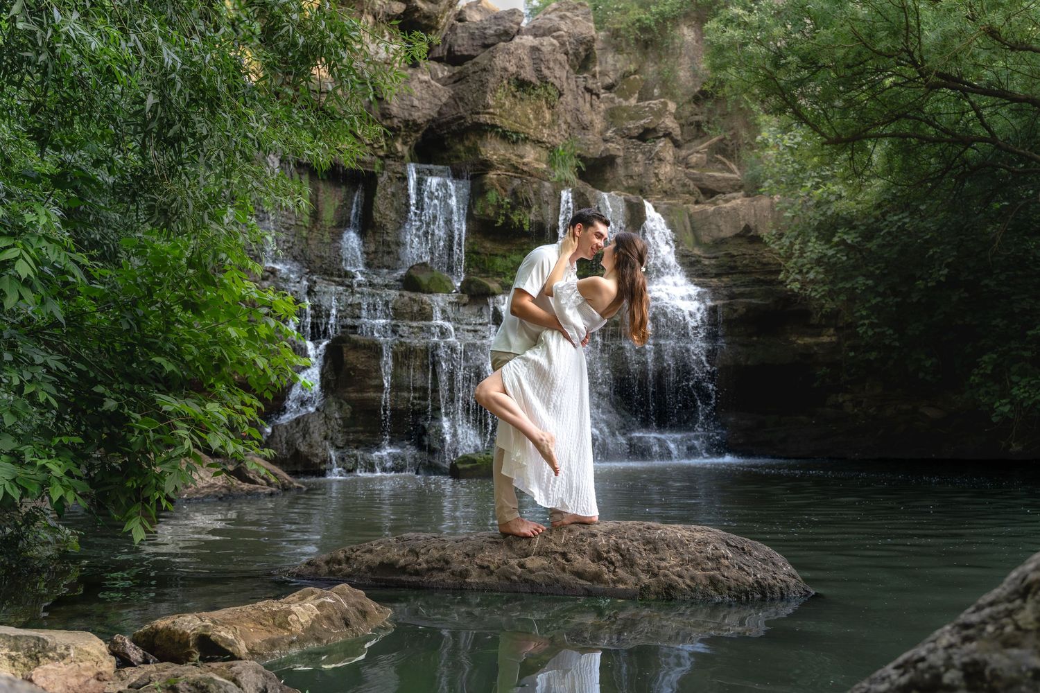 Couple embracing romantically on a rock beside a serene waterfall surrounded by lush green tropical foliage.