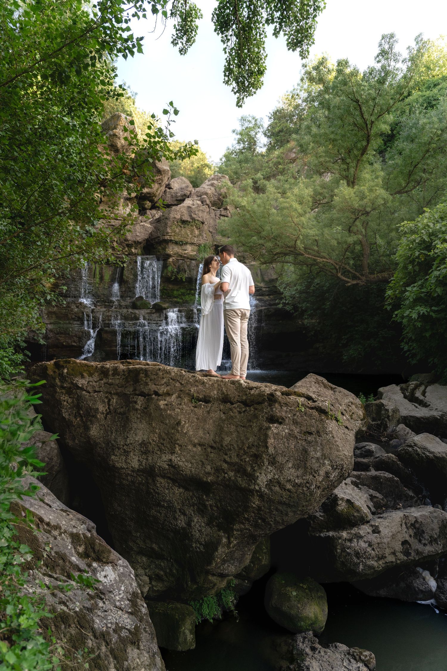Couple in wedding attire standing on rocky ledge before a cascading waterfall surrounded by lush green trees.