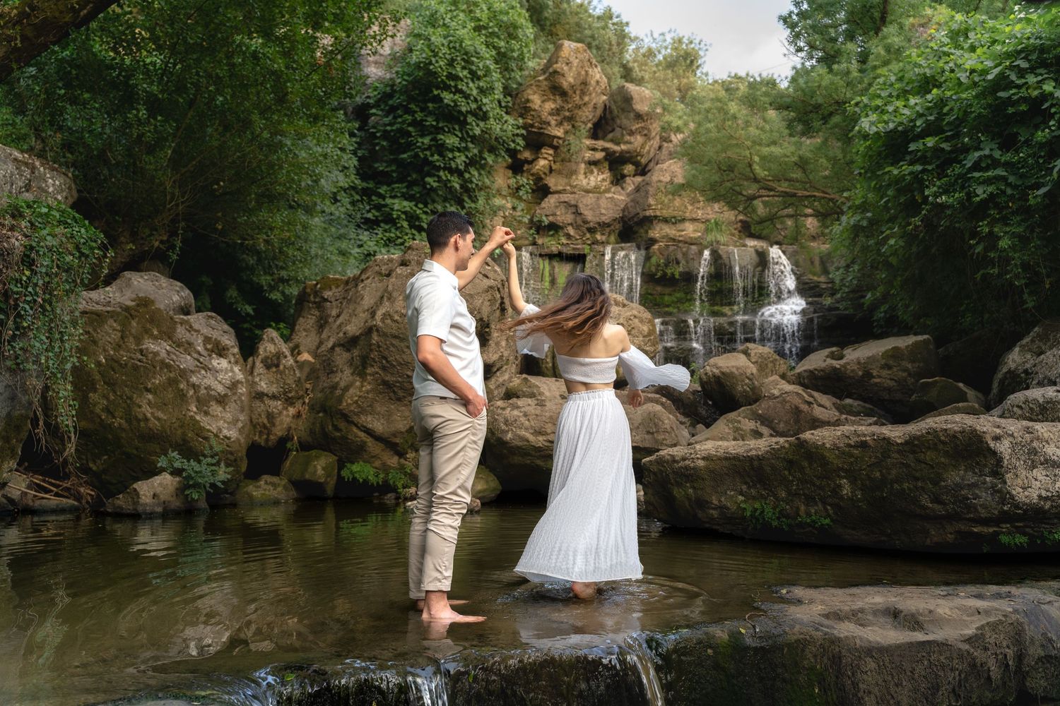 Couple dancing barefoot in a shallow stream with a waterfall and lush green rocks in the background.