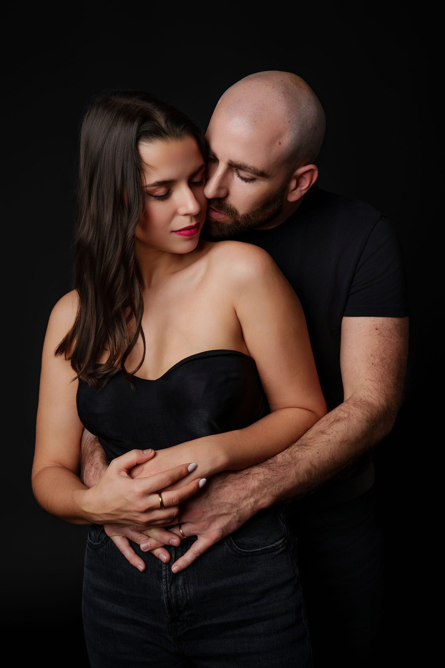 Intimate couple embraces in dark setting wearing black clothing against dramatic black background.