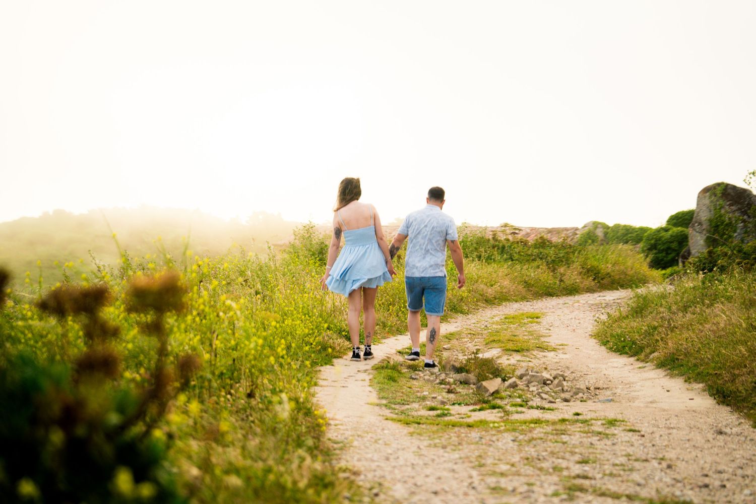 A couple walks along a winding dirt path through a sunny meadow.