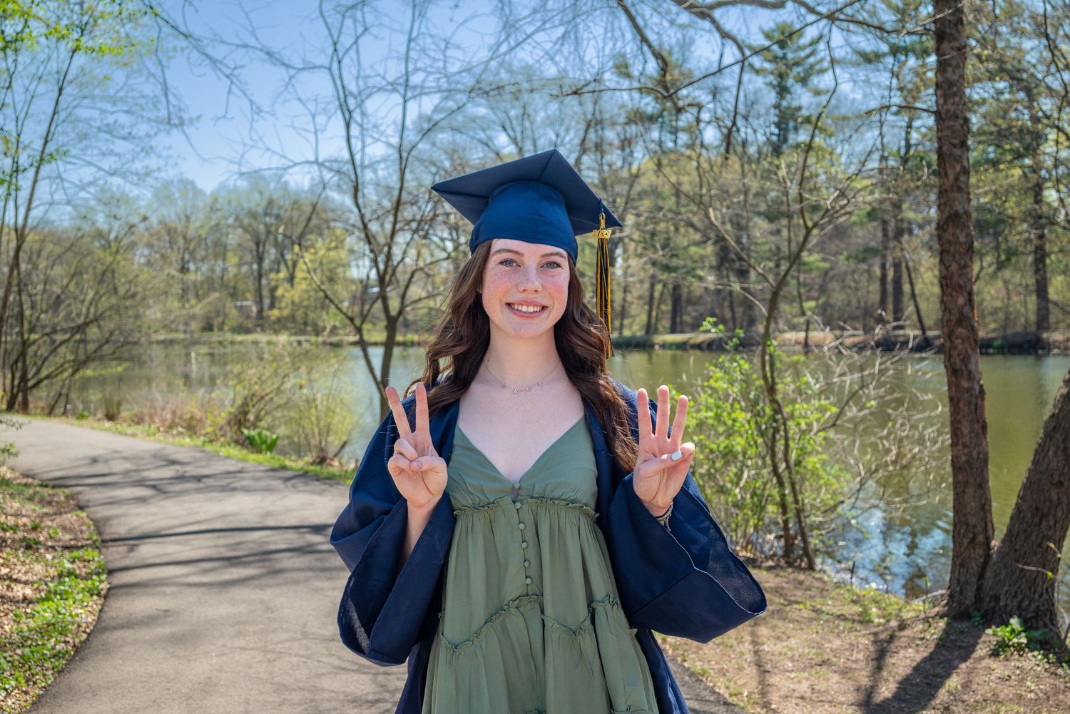 Graduate in cap and gown poses by lakeside path while celebrating on sunny spring day.