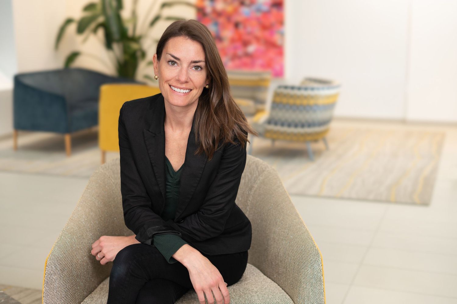 Professional woman in black blazer sits in modern living room with colorful decor.