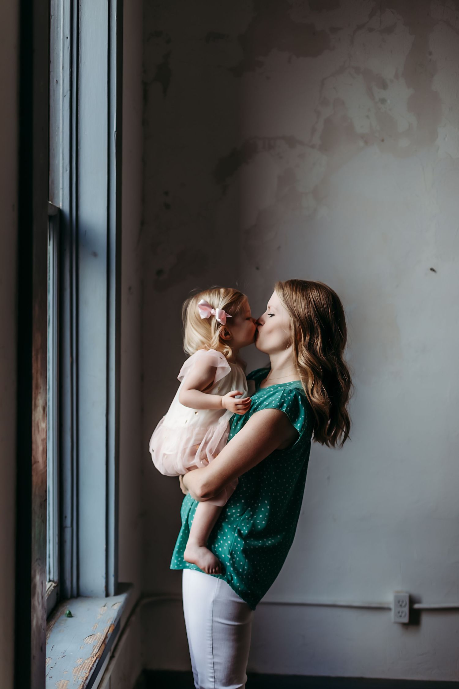Mother and child share tender moment by window in moody indoor setting.