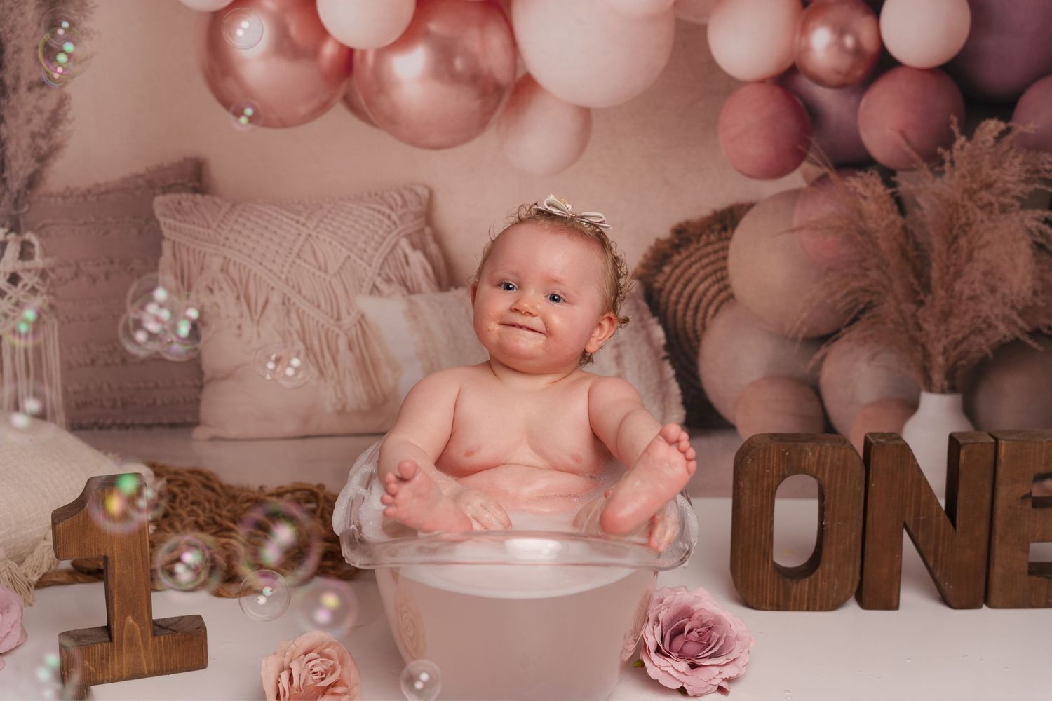 A small baby sits in a pink-themed studio setup with balloons and teddy bears for a first birthday photoshoot.