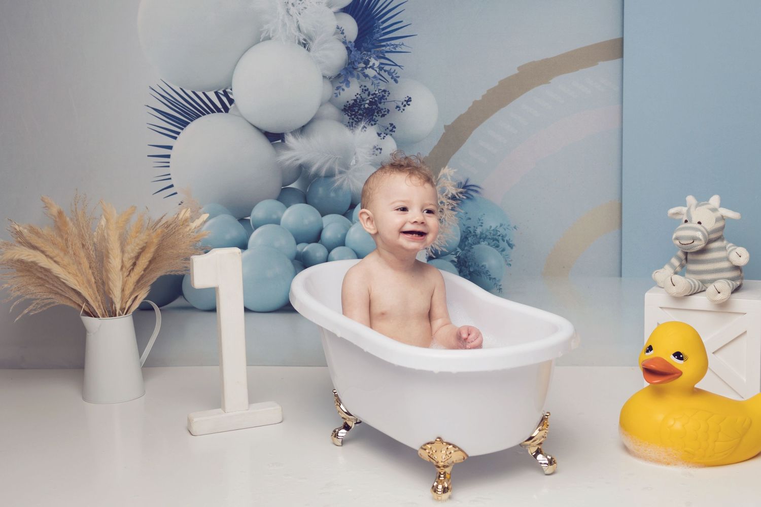 A happy baby sits in a miniature bathtub with a rubber duck against a blue balloon backdrop.