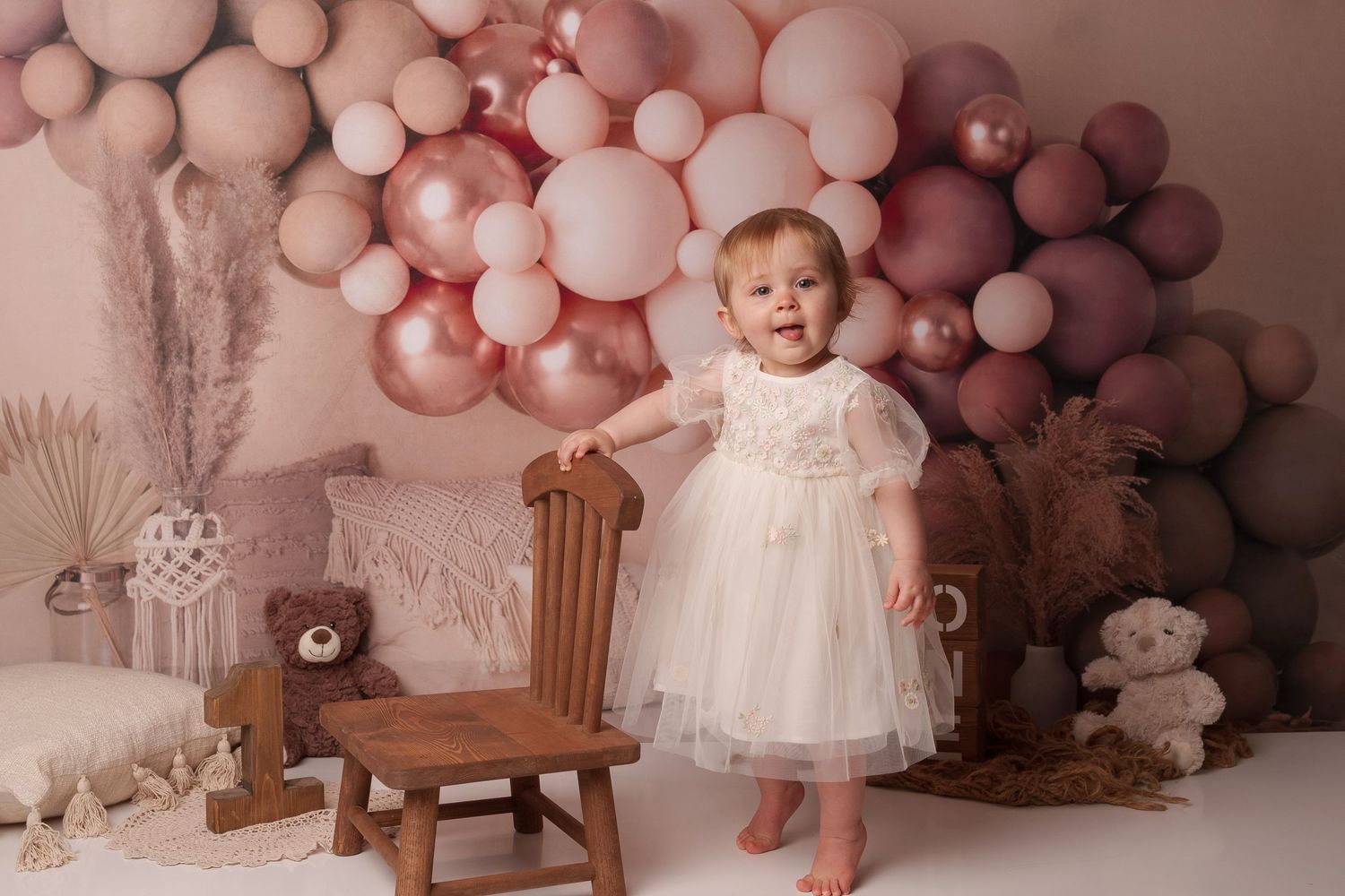 Baby in white dress standing by wooden chair with pink balloon backdrop and teddy bears.