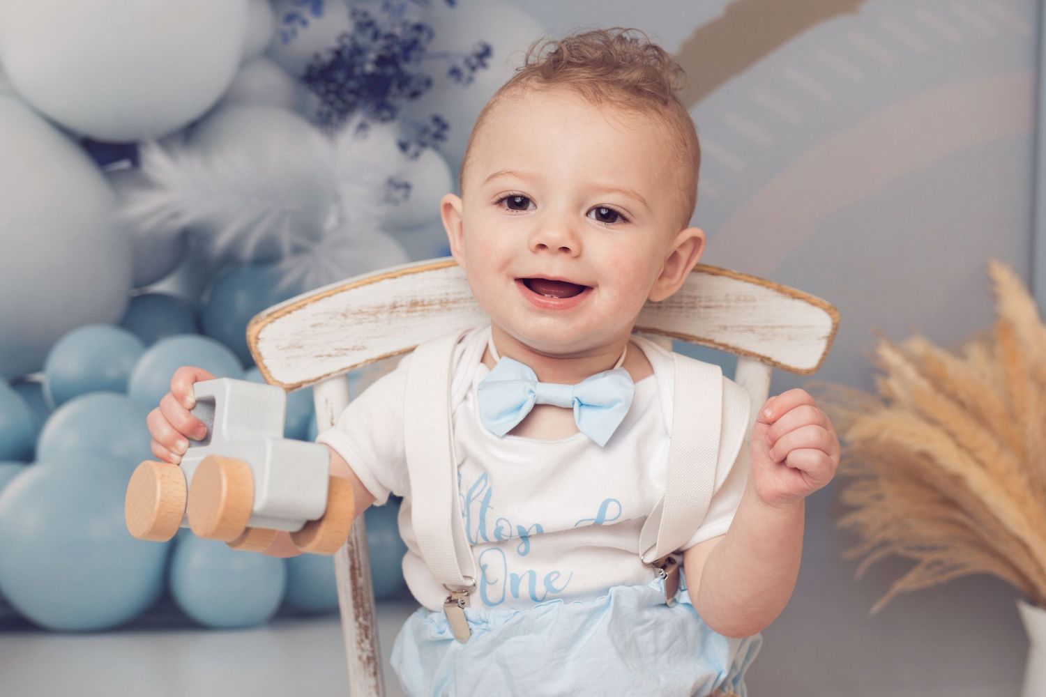 A smiling baby in light blue outfit sits in a decorated studio with blue balloons and wheat stalks.