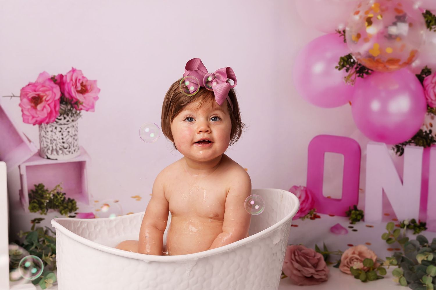 Baby in vintage-style white bathtub with pink balloon and floral decorations for first birthday shoot.