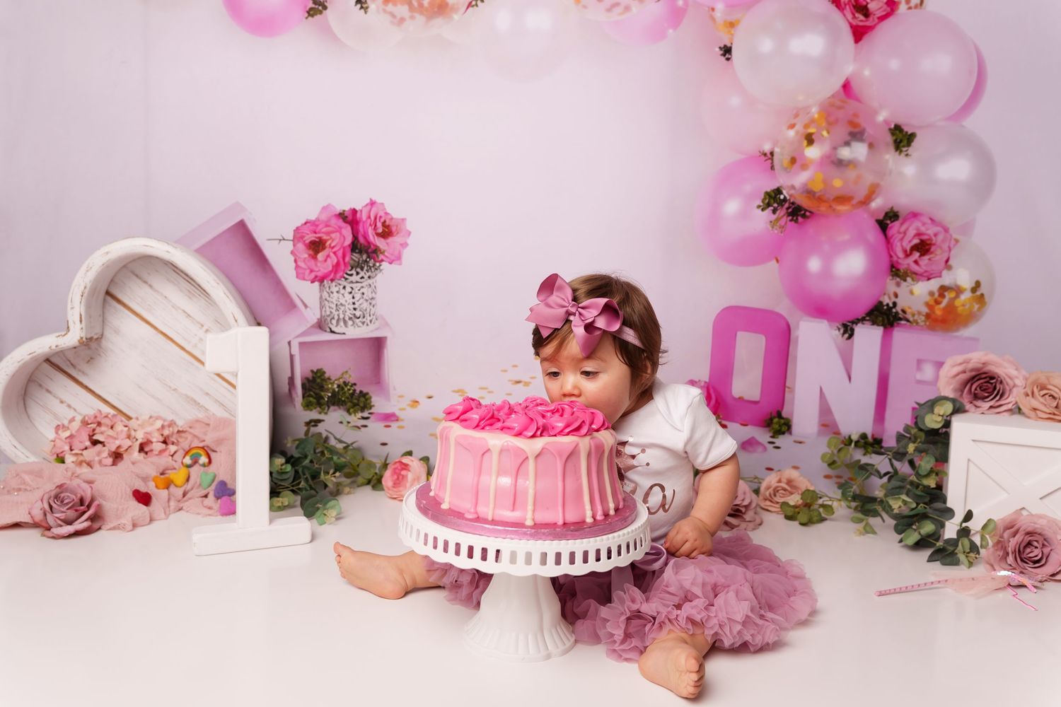 Pink first birthday cake smash setup with balloons, roses and decorative heart frame against light pink backdrop.